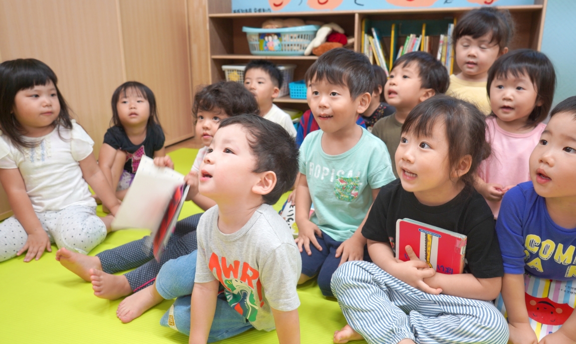 読み聞かせに集中する子供達。子供達の後ろには、子供達が描いた絵が並んでいます。