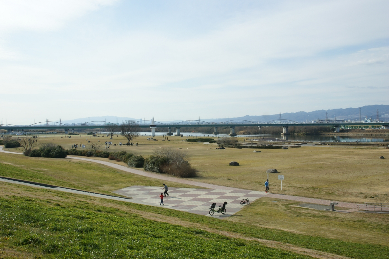 枚方河川公園の景色。青空が広がり、公園では人々がスポーツをしています。奥には、ひらかたパークの観覧車が見えます。